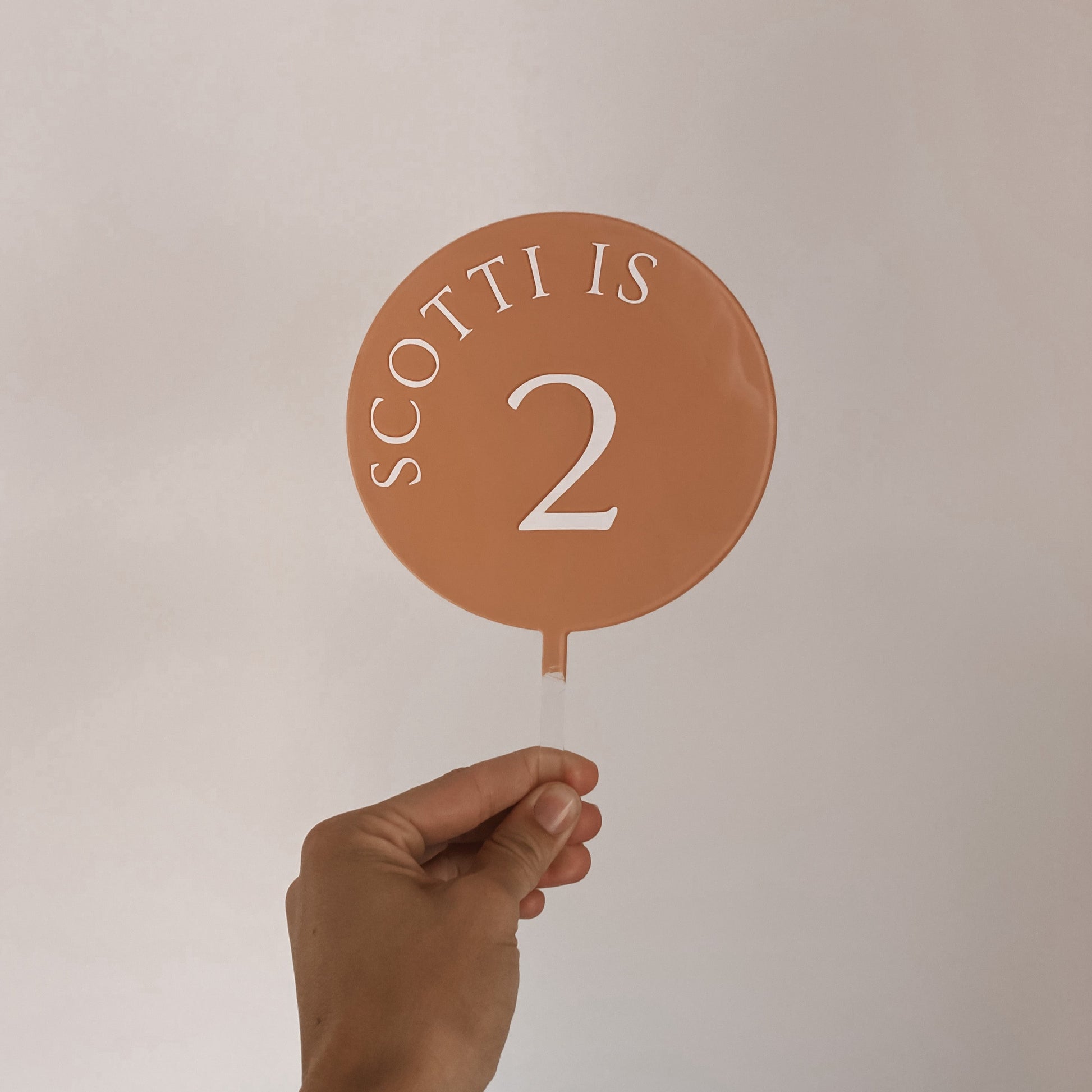 5 inch round cake topper in coral pink, with white print font that says "Scotti is 2"
